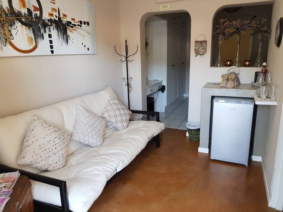 Un espacio habitable compacto con sofá blanco, almohadones estampados, un refrigerador pequeño, un perchero y un espejo con detalles decorativos.