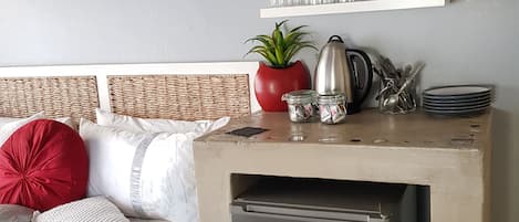 A compact bedroom with a bed, a small kitchenette, a clock, and a shelf with glassware.