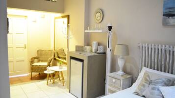 Un dormitorio con cama, mesita de noche, lámpara y un refrigerador pequeño. En la esquina, hay una silla y una mesa con una planta.