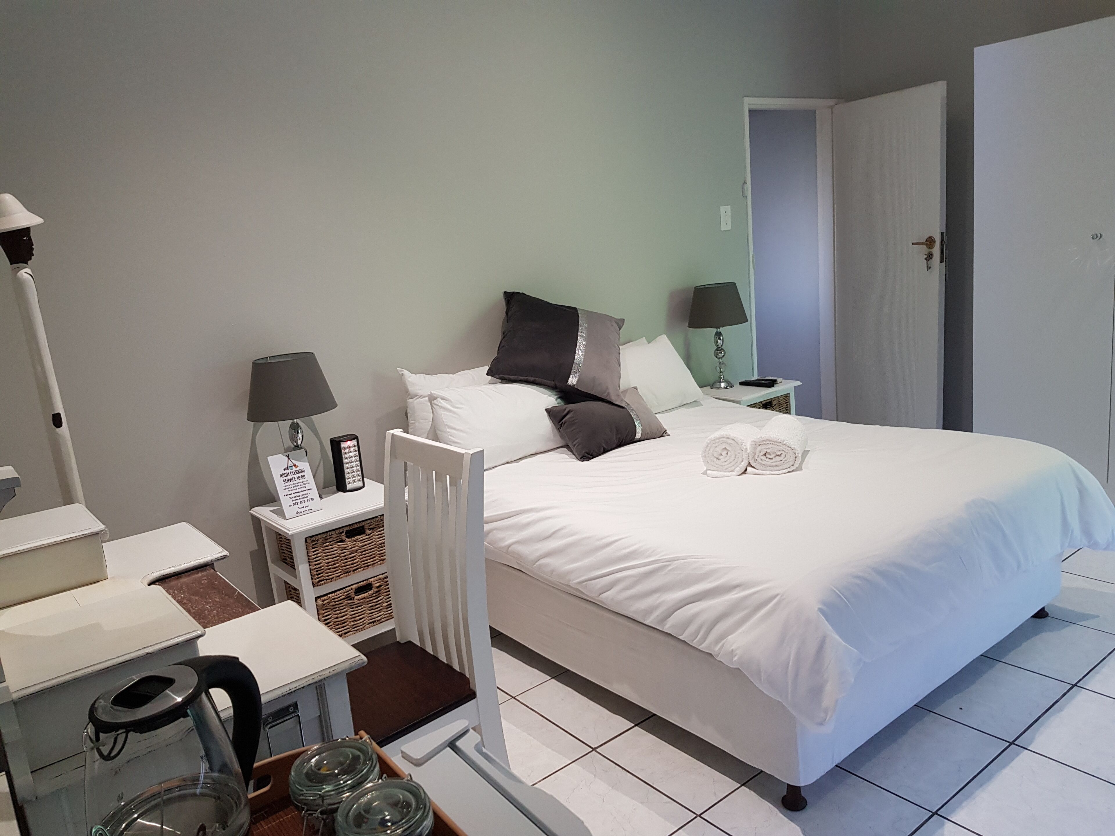 A neatly arranged hotel room with a bed, bedside tables, a chair, a lamp, and a kettle.