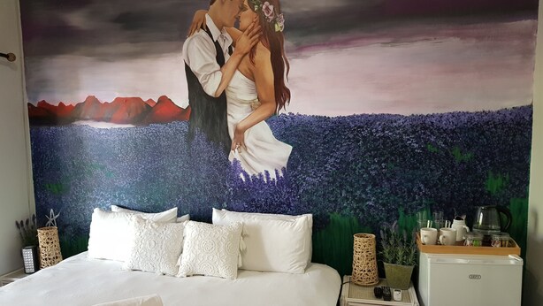 Quarto com cama grande, roupa de cama branca, almofadas decorativas, mesa de cabeceira pequena com abajur e tapeçaria na parede retratando um casal.