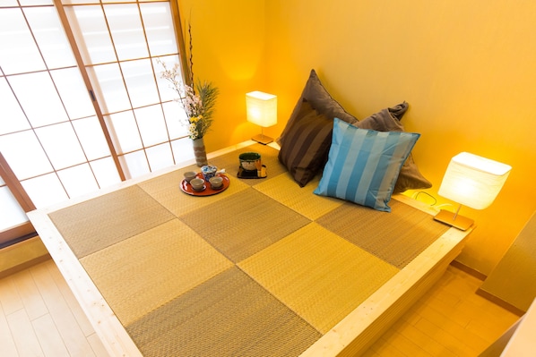 A tatami mat room with a low bed, two pillows, a small table with cups, and a lamp on the side.