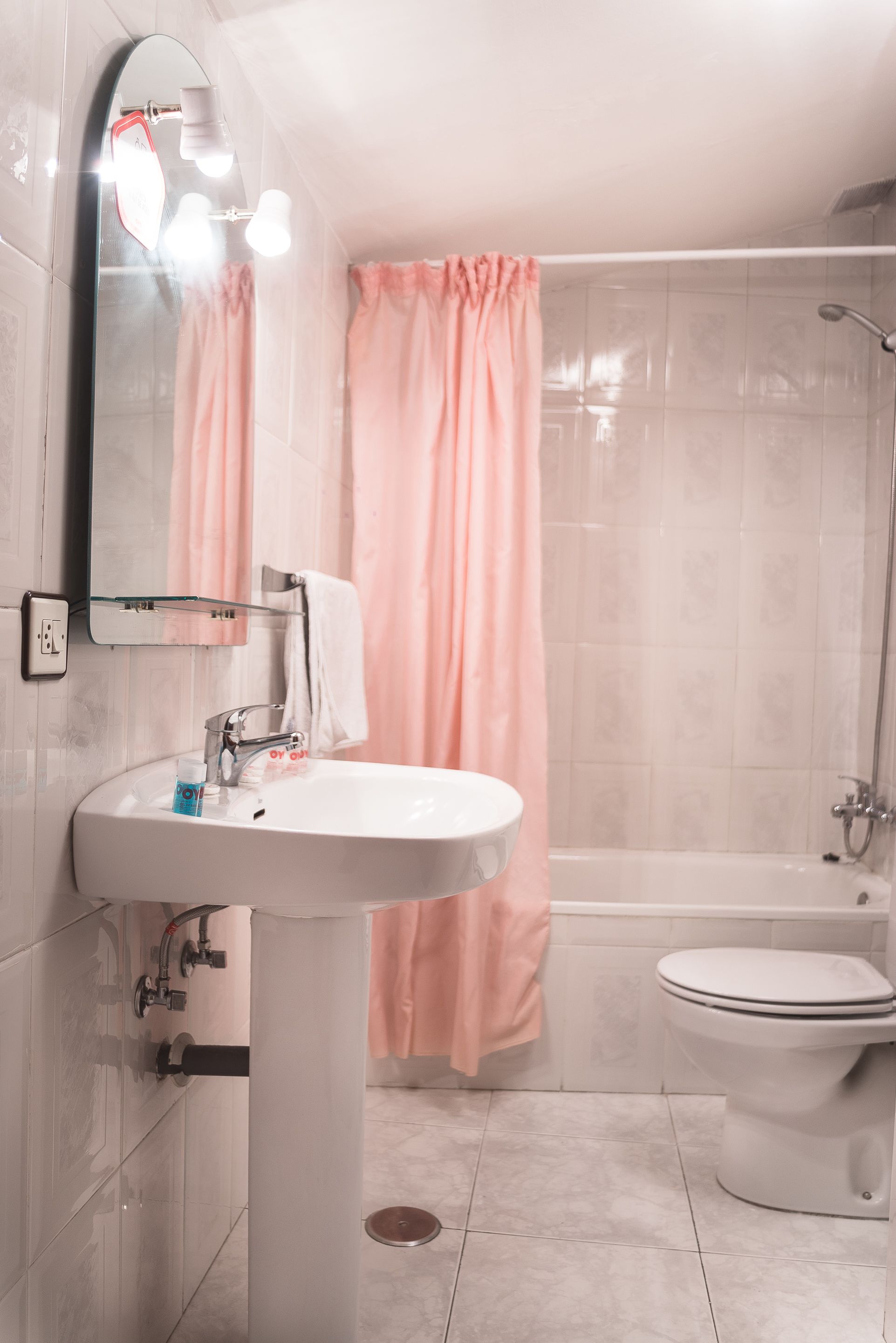 Un baño con lavamanos blanco, espejo e inodoro. Hay una zona de ducha con cortina rosa.