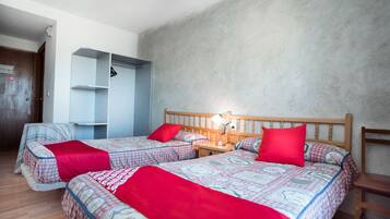 A bedroom with two beds, each with a red blanket and patterned bedding. There is a wooden headboard, a bedside table with a lamp, and a wooden chair.