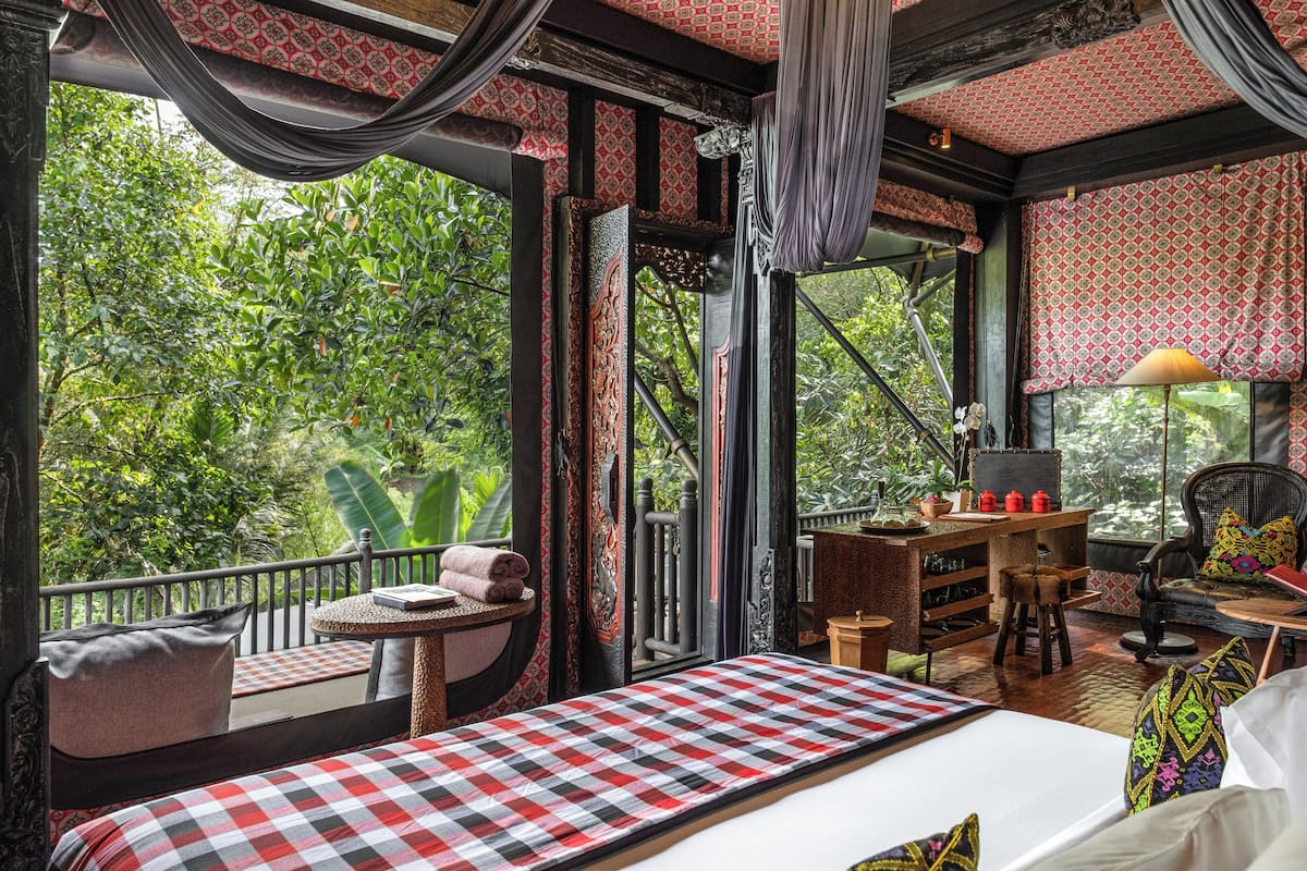 Ein Zimmer mit kariertem Bett, einem kleinen Tisch mit Lampe, einem Stuhl und Blick auf üppiges Grün draußen.
