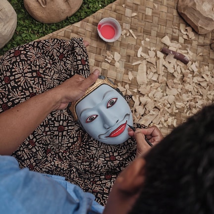 A person painting a mask on a woven mat with wood shavings around.