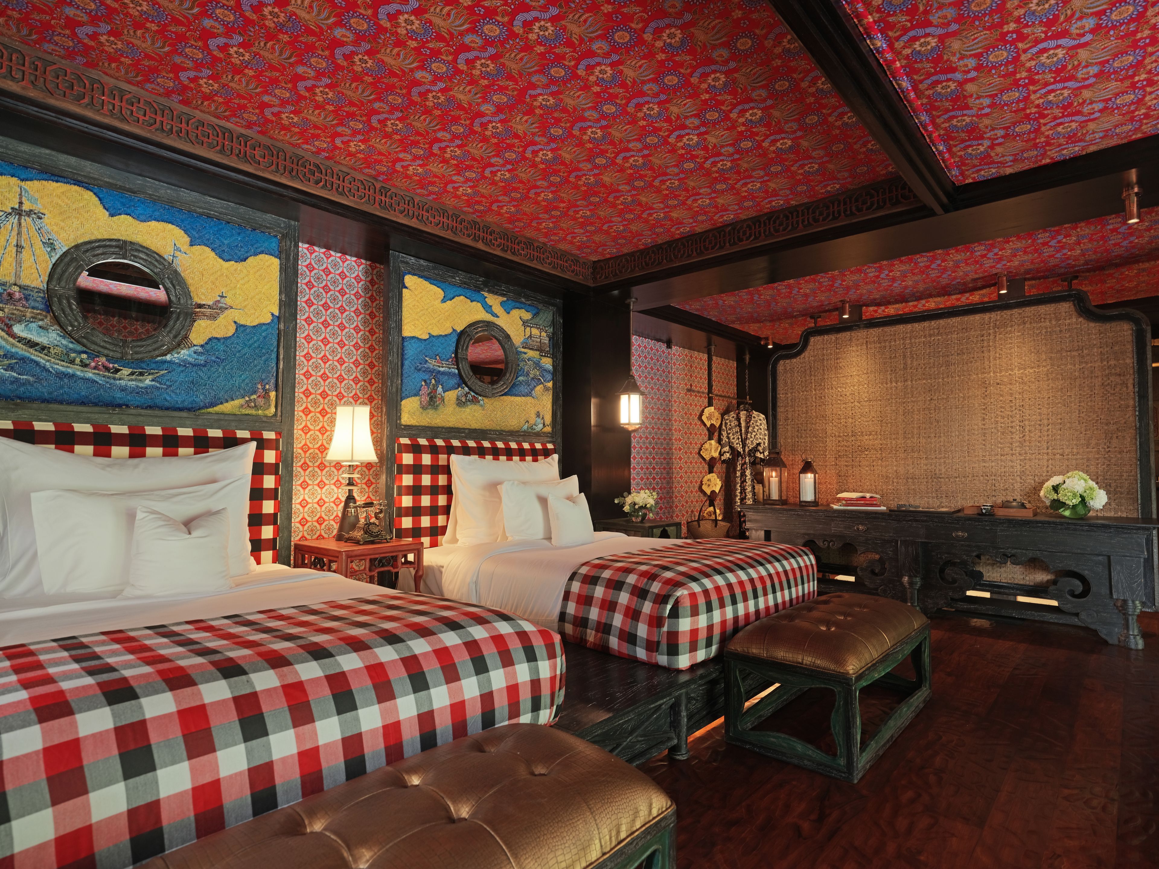 Ein Hotelzimmer mit zwei Betten, einer karierten Tagesdecke, einer hölzernen Bank, einem Nachttisch mit Lampe sowie einer Wand mit einem runden Spiegel und Kunstwerken.