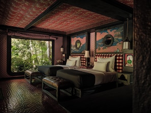 A room with two beds, a large window with a view of greenery, patterned wallpaper, and a decorative ceiling.