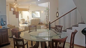 Een eetgedeelte met een glazen tafel, omringd door houten stoelen, een trap met een leuning en een woonkamer op de achtergrond.