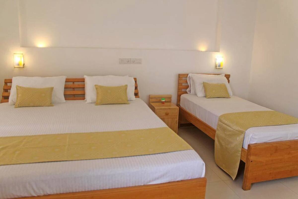 Een hotelkamer met twee enkele bedden, elk met een houten hoofdeinde, wit beddengoed en een gele plaid. Er staan nachtkastjes met lampen en een klein nachtkastje met een klok.