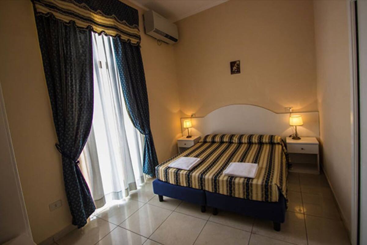 A bedroom with a bed, two bedside tables, a wall-mounted air conditioner, and a window with curtains.