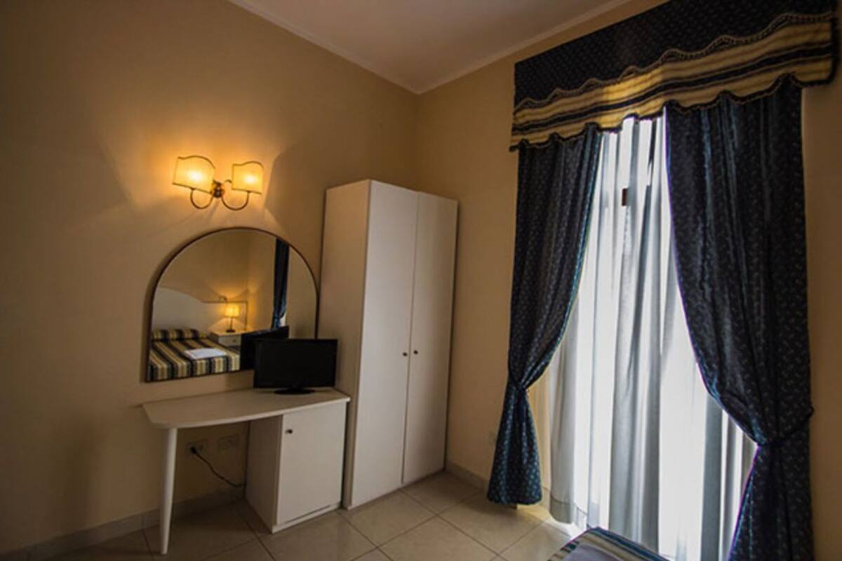 A room with a desk, a television, a wardrobe, a mirror, and a window with curtains.