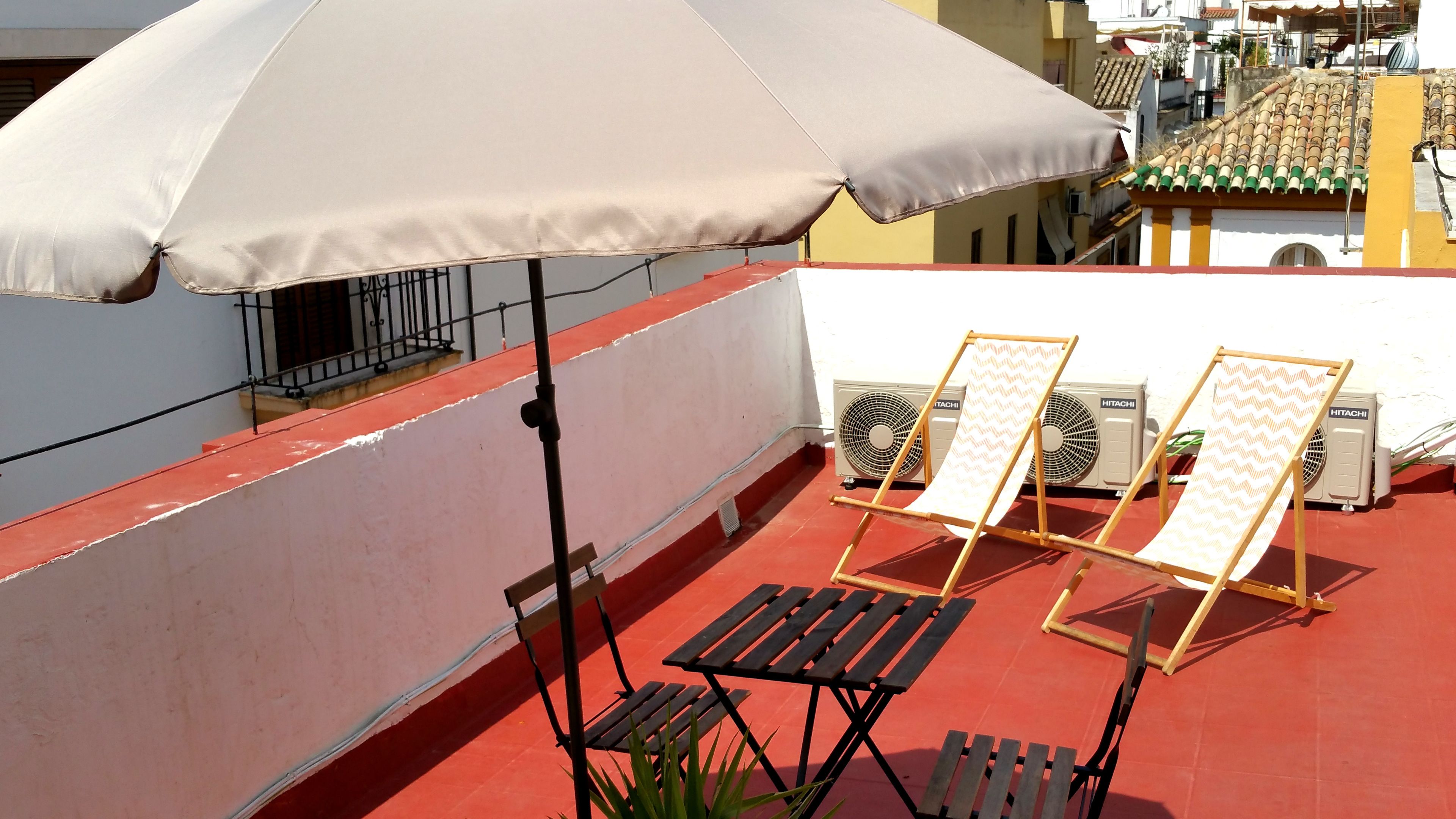 A rooftop terrace with a beige umbrella, two wooden lounge chairs, and two air conditioning units.