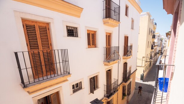 A narrow street with multi-story buildings, balconies with wooden shutters, and a person walking.