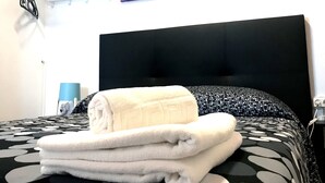 A neatly made bed with a black headboard and a patterned bedspread, a stack of white towels, and a colorful sign above the bed advertising ARC HOUSE.