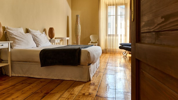 Un dormitorio con piso de madera, una cama con ropa blanca, una mesita de noche, una lámpara y una silla.