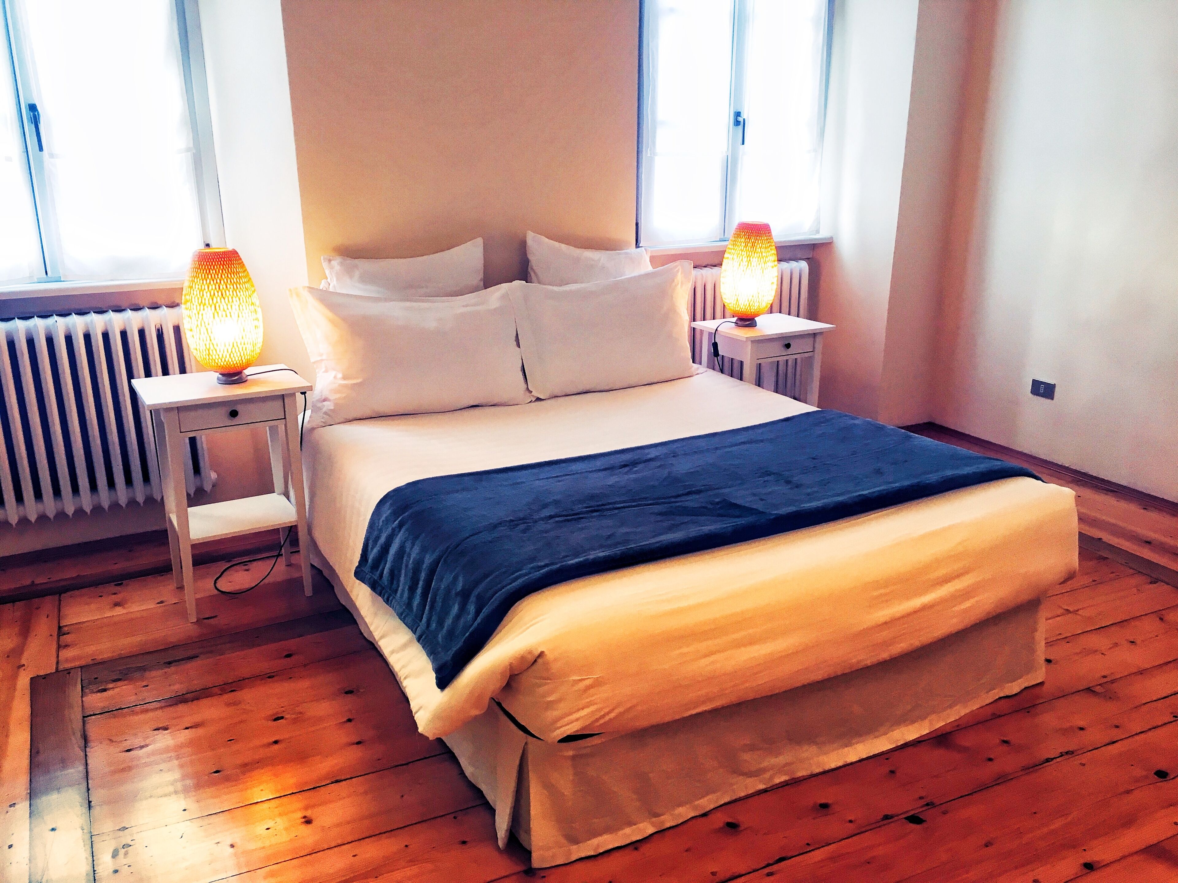 A neatly made bed with white and blue bedding, two bedside lamps, and a radiator in a room with wooden flooring.