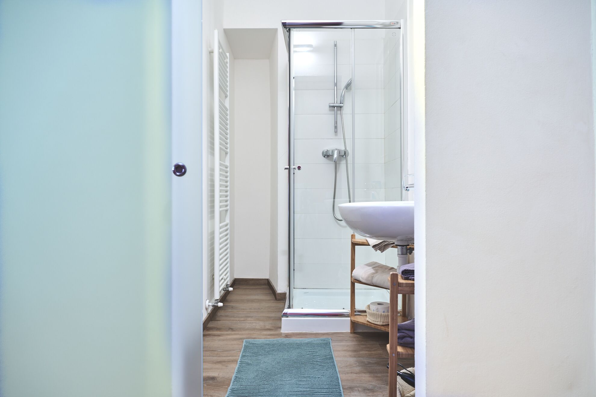 A narrow bathroom with a shower, a sink, and a wooden shelf with towels.
