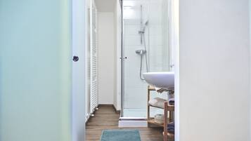 A narrow bathroom with a shower, a sink, and a wooden shelf with towels.