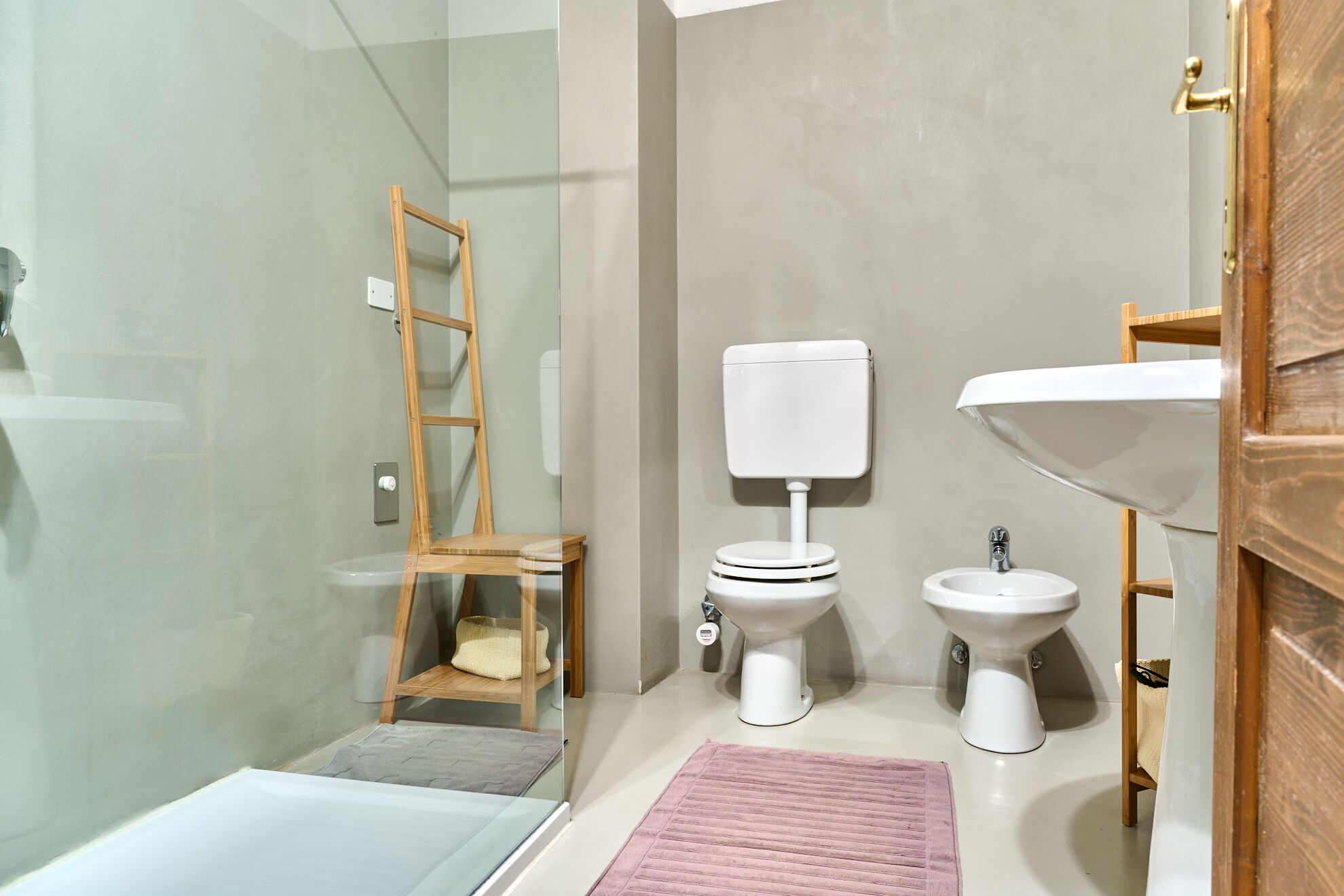 A bathroom with a glass shower enclosure, a wooden ladder, a toilet, a bidet, and a sink.