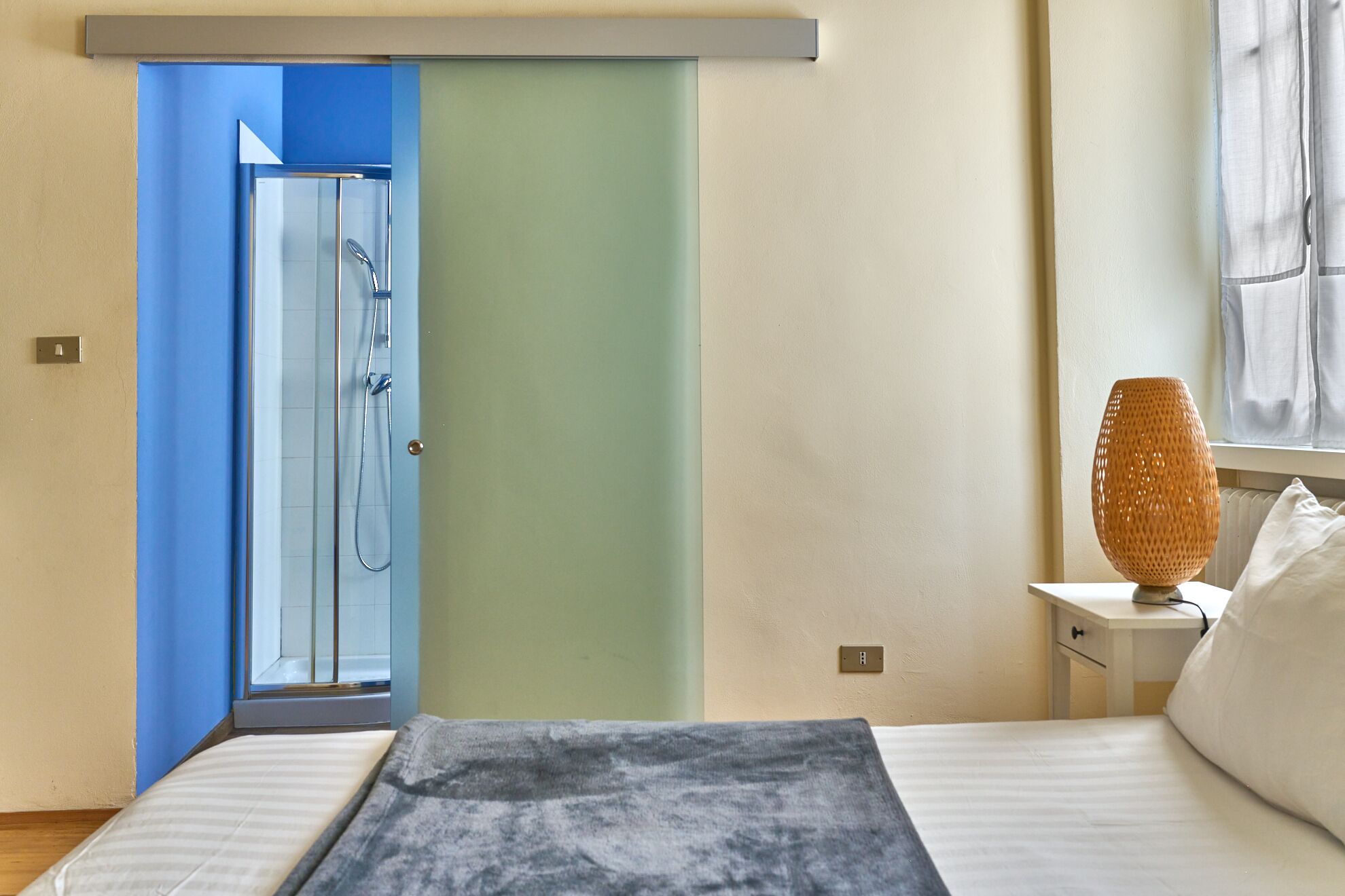 A bedroom with a bed, a nightstand, a lamp, and a bathroom visible through a door.