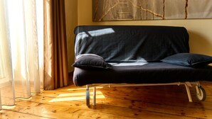 Una habitación con piso de madera, un sofá negro con dos cojines, una silla blanca y un cuadro abstracto grande en la pared.
