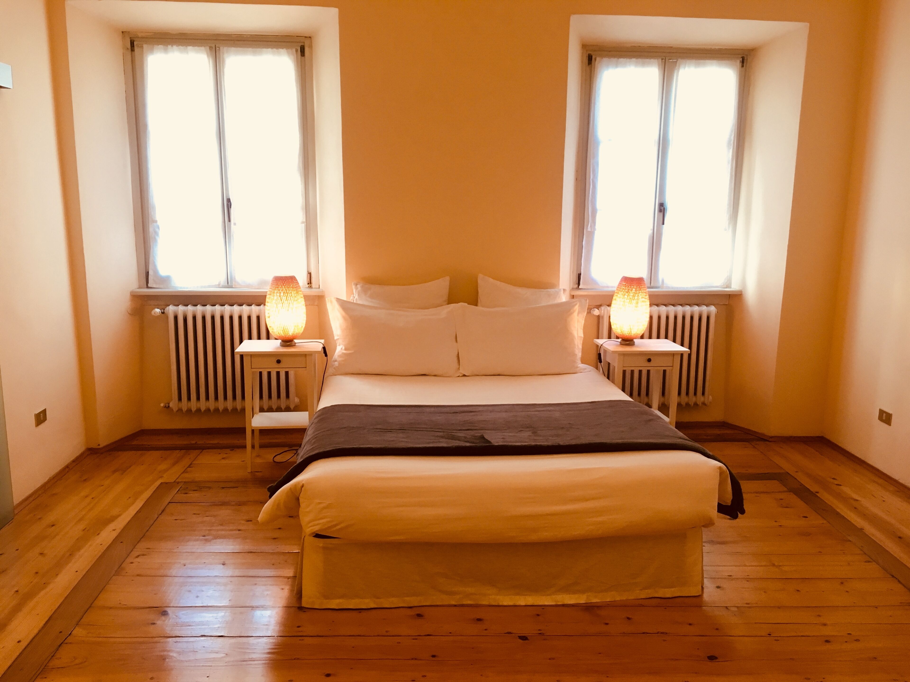 A bedroom with a bed, two bedside tables, a radiator, and two windows with curtains.