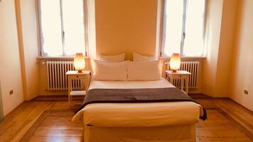 A bedroom with a bed, two bedside tables, a radiator, and two windows with curtains.