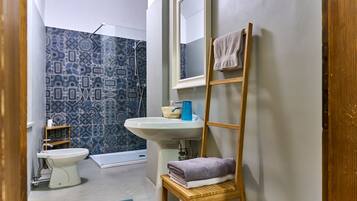 Une salle de bain équipée de toilettes, d’un lavabo et d’une douche. On y trouve un porte-serviettes avec des serviettes et une étagère en bois avec un panier.