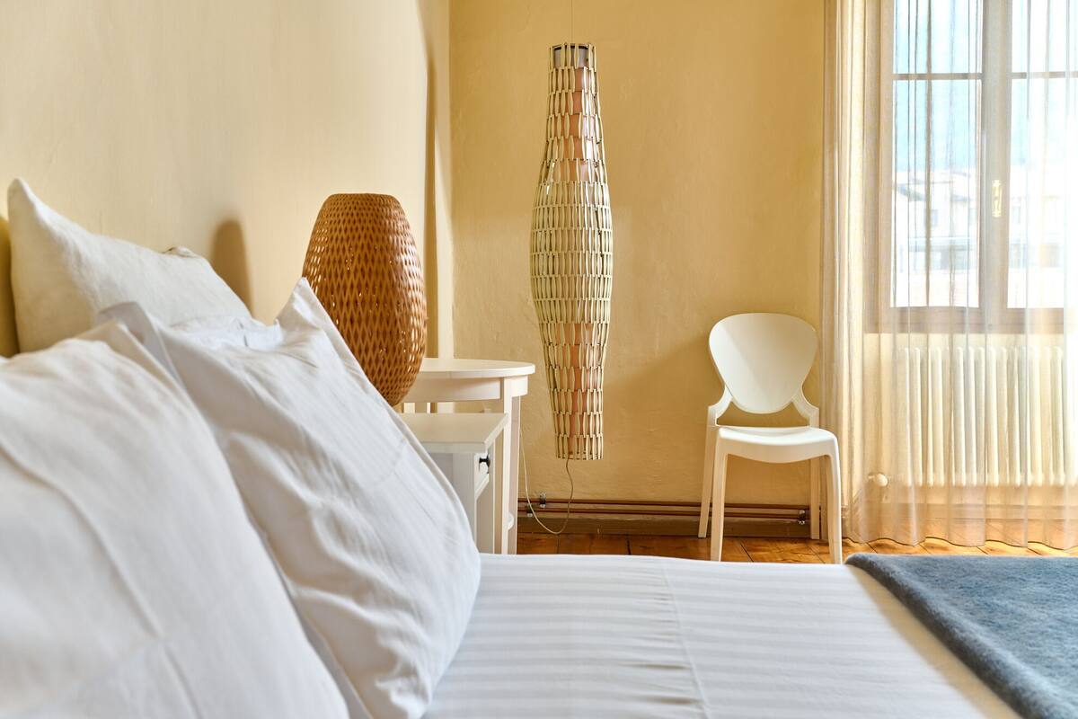 A neatly made bed with white linens, a white chair, and a hanging light fixture in a room with a large window and a view of the city.