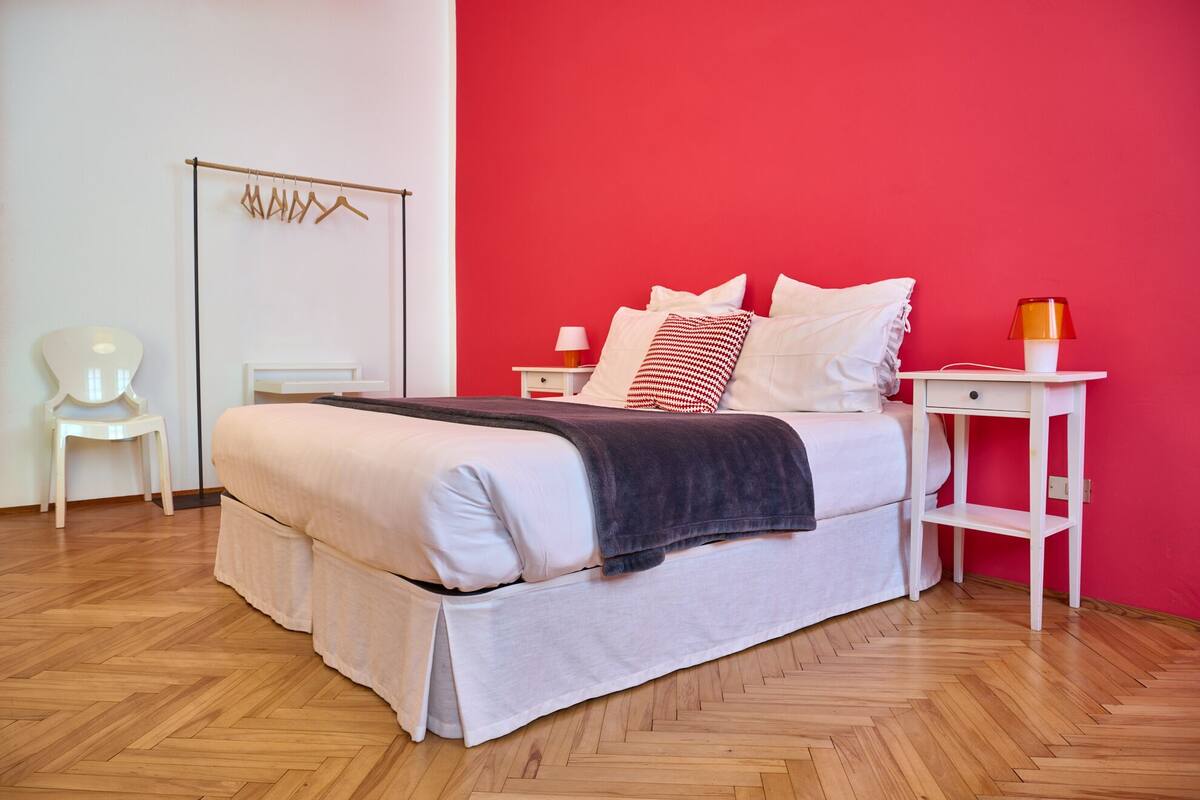 A bedroom with a bed, a bedside table, a chair, and a wall with a red accent.
