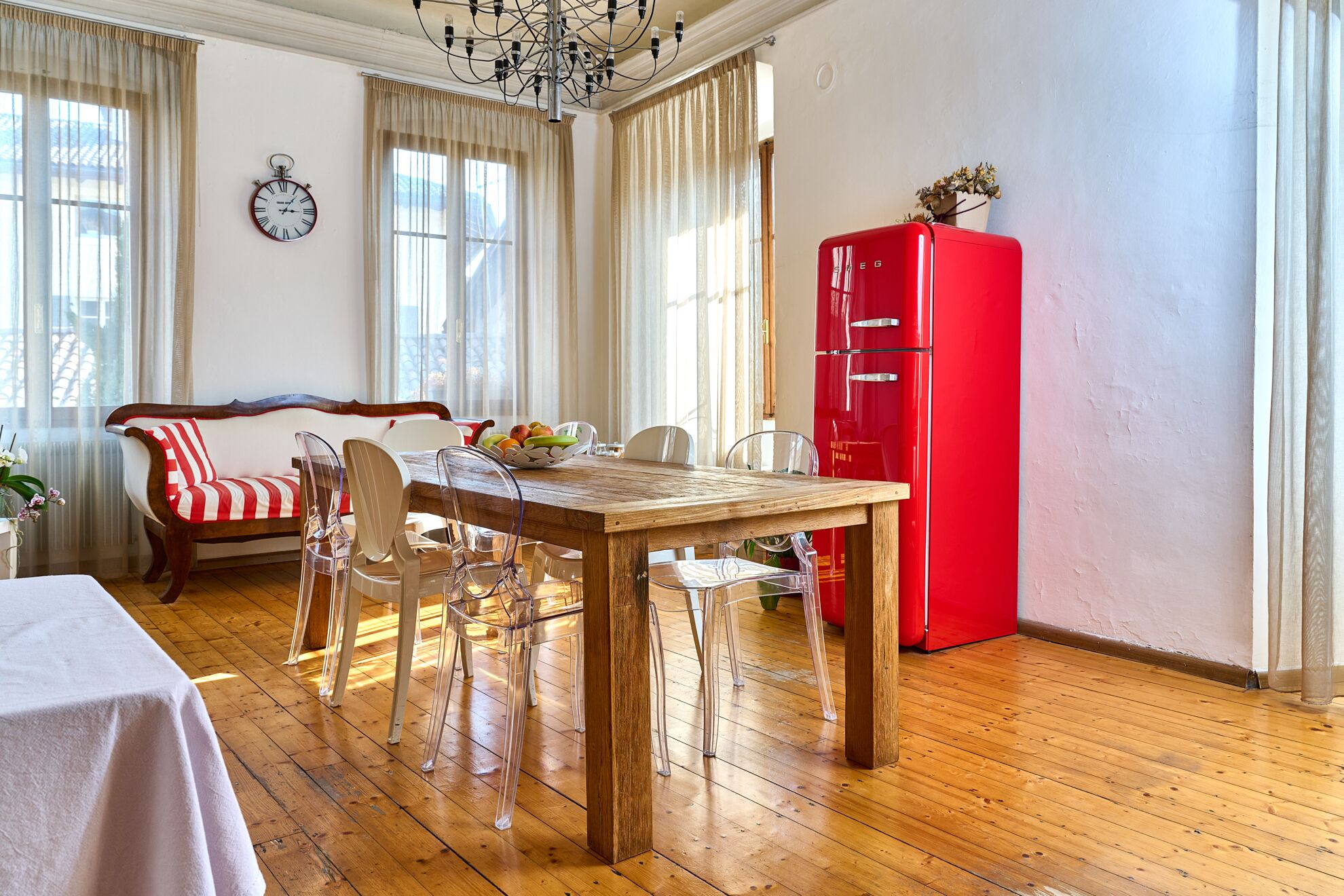 A dining area with a wooden table, clear chairs, a red refrigerator, and a sofa with striped cushions.