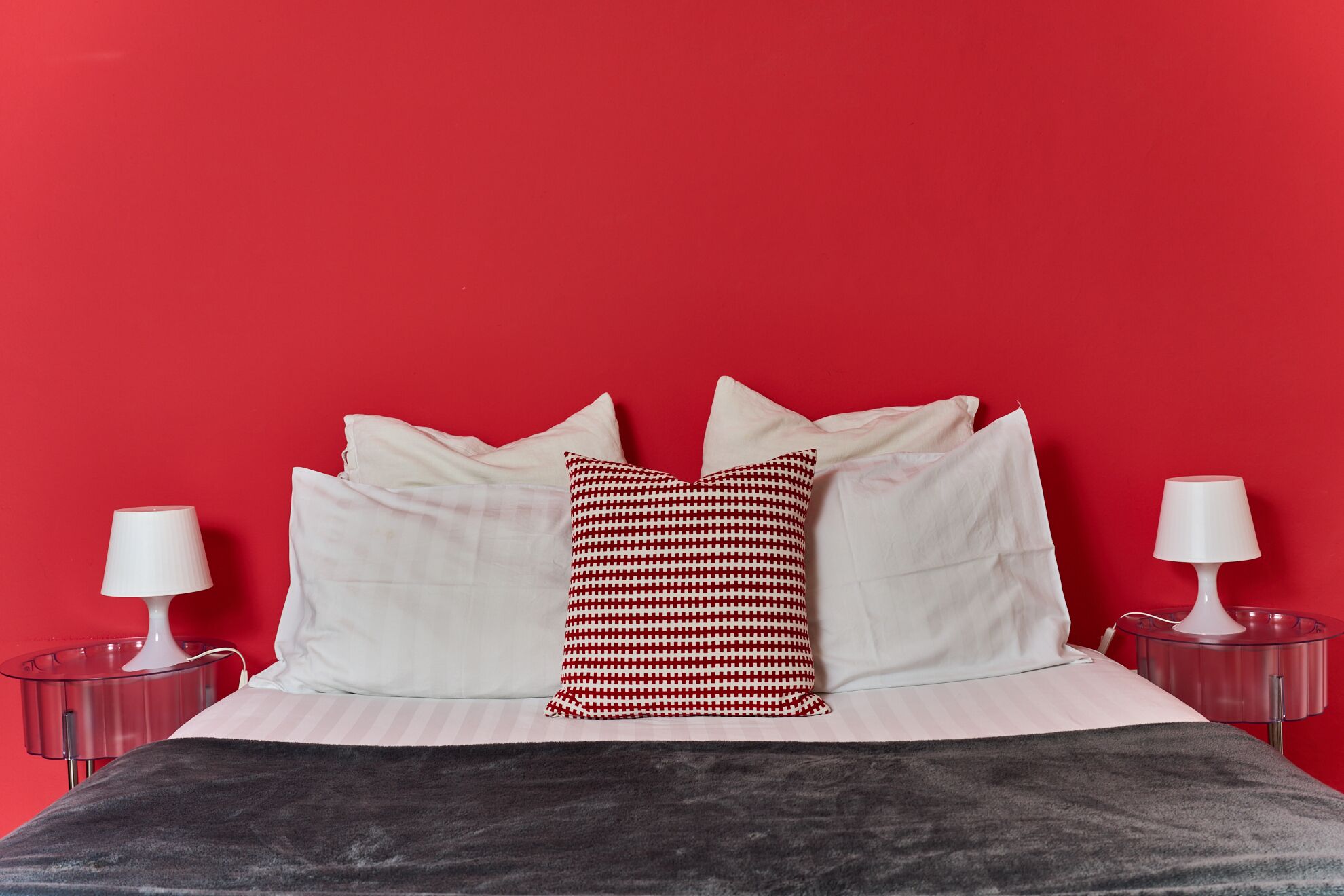 Una cama con almohadas blancas y una almohada con estampado rojo y blanco, frente a una pared roja. Hay dos mesitas de noche con lámparas.