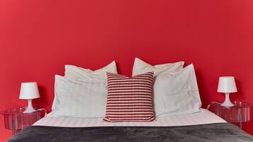 Una cama con almohadas blancas y una almohada con estampado rojo y blanco, frente a una pared roja. Hay dos mesitas de noche con lámparas.