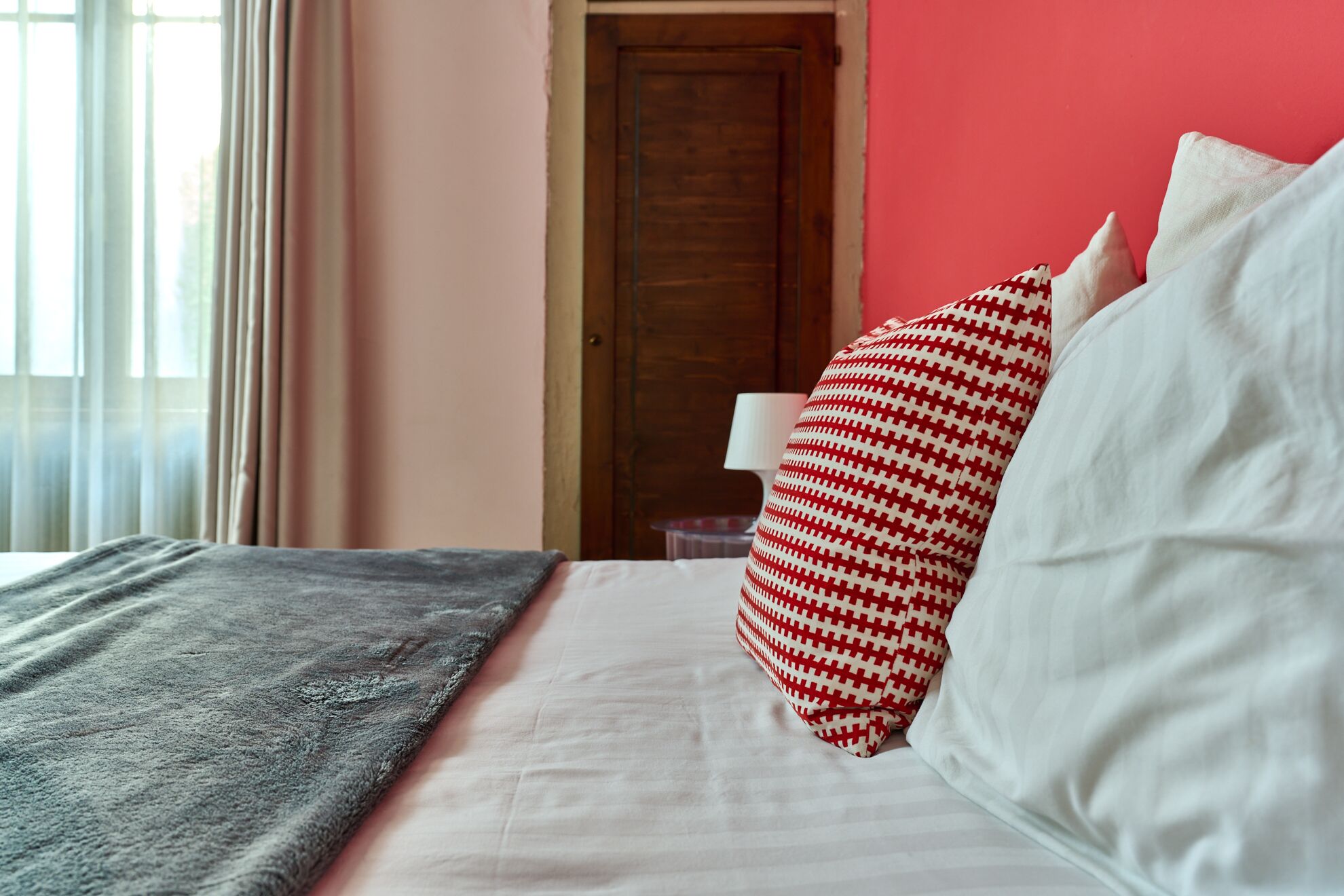 A neatly made bed with a grey blanket, white sheets, and a red and white patterned pillow.
