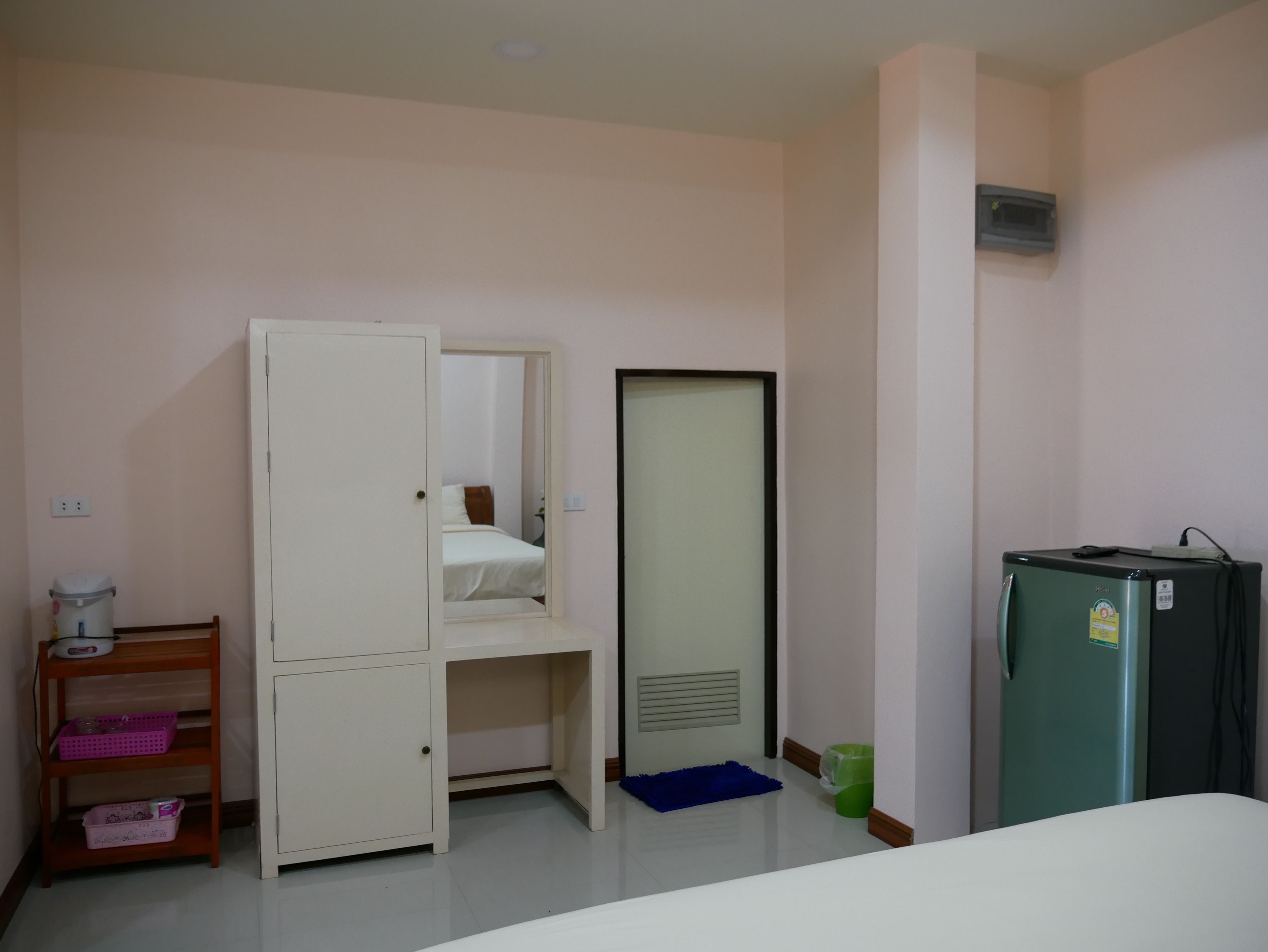 A hotel room with a bed, a wardrobe, a small desk with a lamp, a refrigerator, and a trash bin.