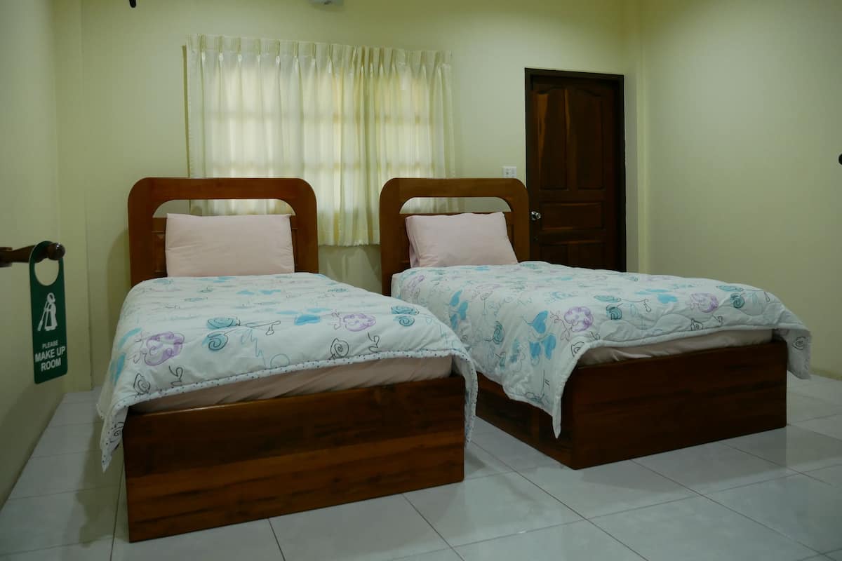 A room with two wooden beds, each with a white bedspread featuring blue and purple floral patterns. There is a wooden door and a window with sheer curtains.
