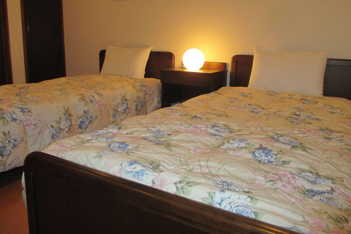 Una habitación con dos camas individuales, cada una con cabecero de madera y colcha estampada. Entre las camas hay una mesita de noche con una lámpara.