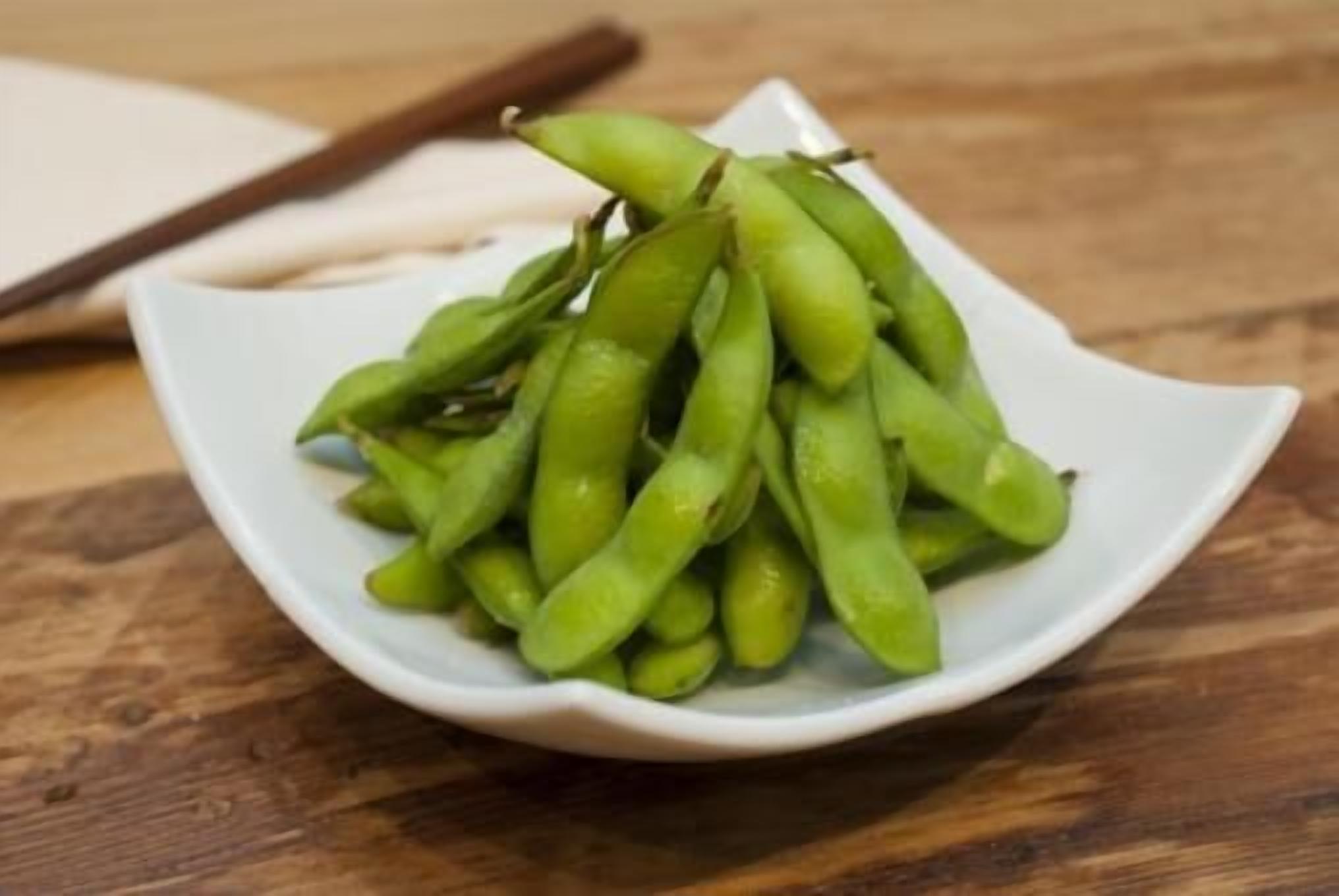 A white bowl filled with green edamame on a wooden surface.