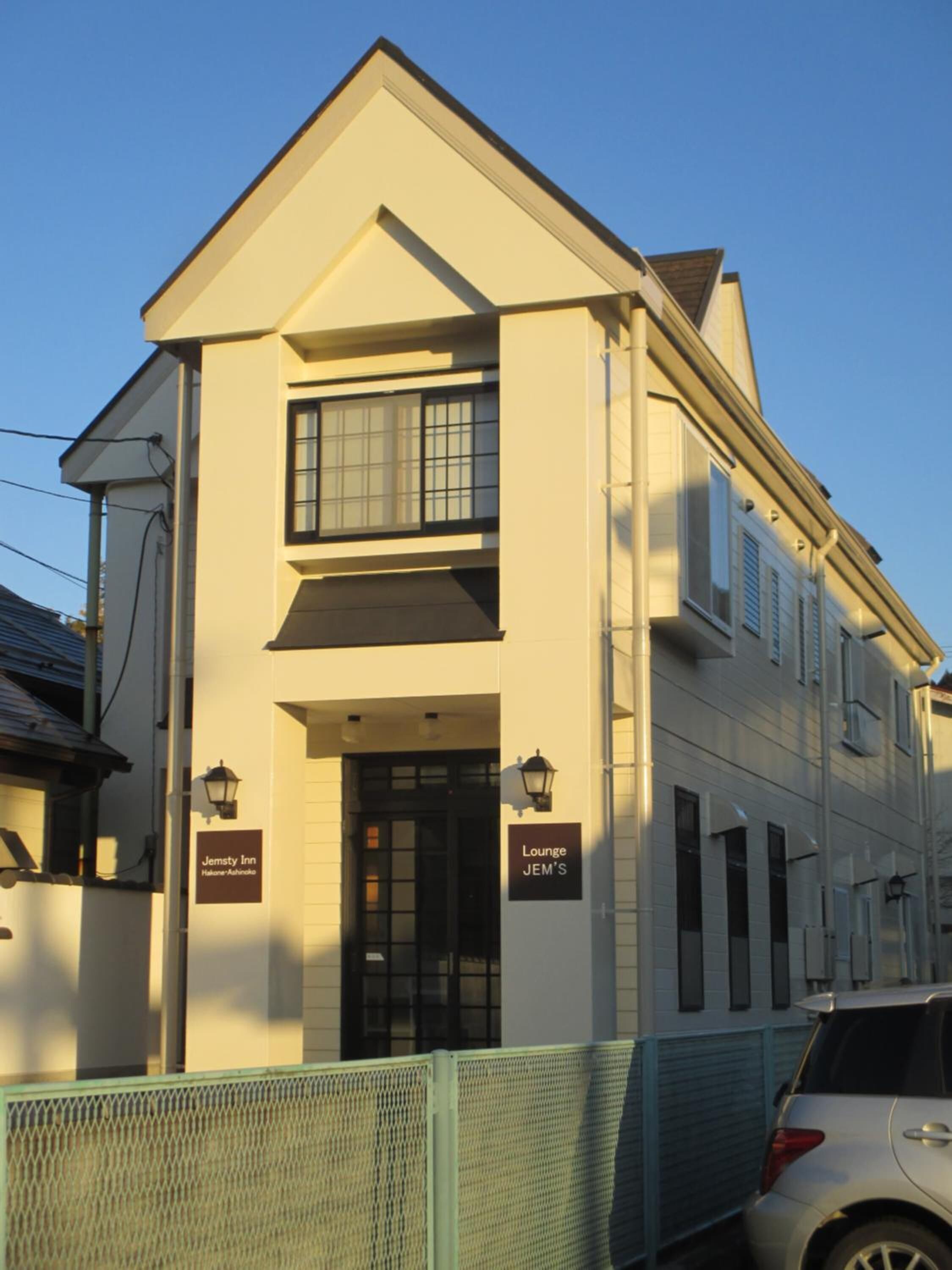 Un bâtiment à deux étages, doté d’une façade blanche, d’une entrée encadrée de noir et de deux enseignes indiquant « Jemsty Inn » et « Lounge JEM’S ».