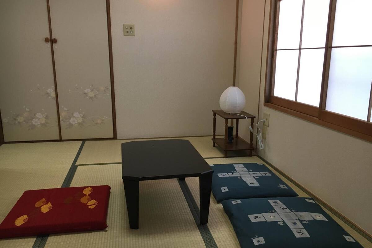 Una habitación con esteras de tatami, una mesa negra, cojines rojos y azules, y un farol de papel sobre un soporte de madera.