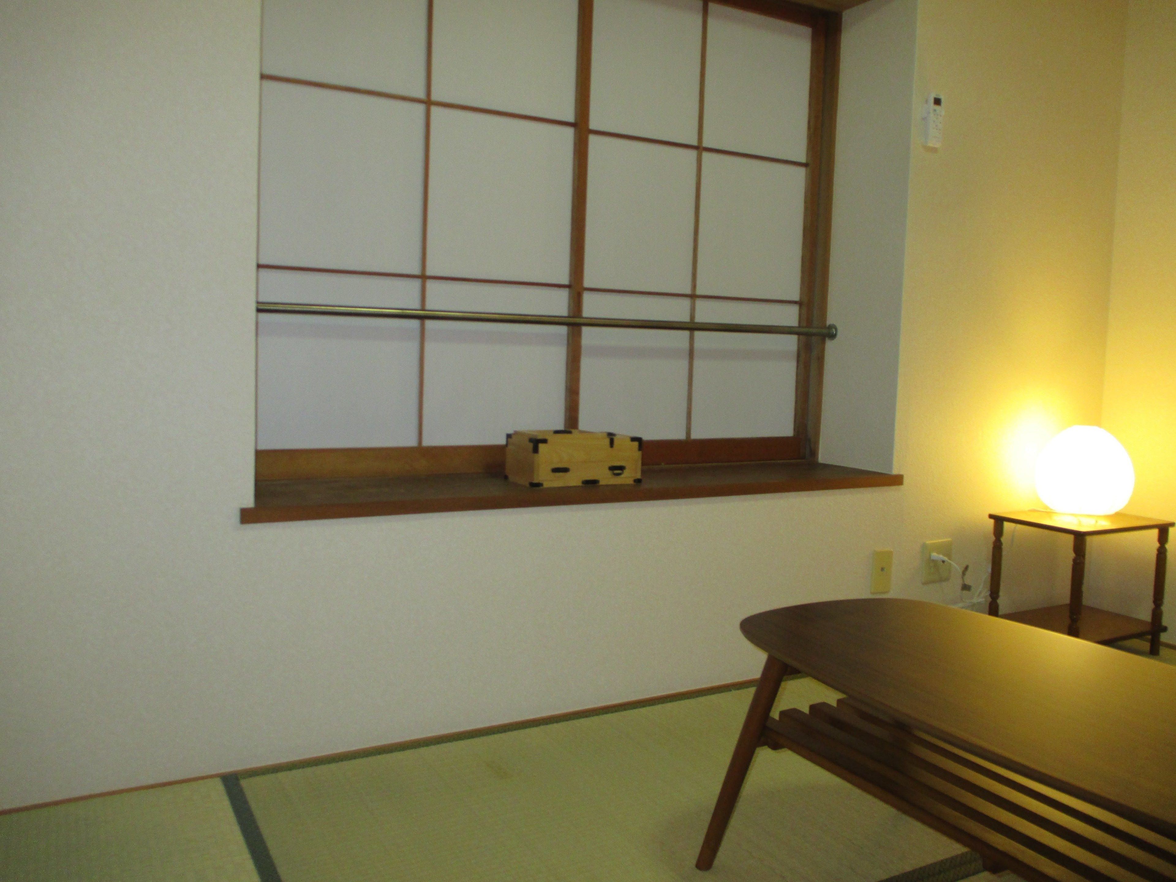 Une pièce au sol recouvert de tatamis, une petite table basse en bois, une fenêtre avec un cadre en bois et une petite table sur laquelle se trouve une lampe qui diffuse une lumière douce.