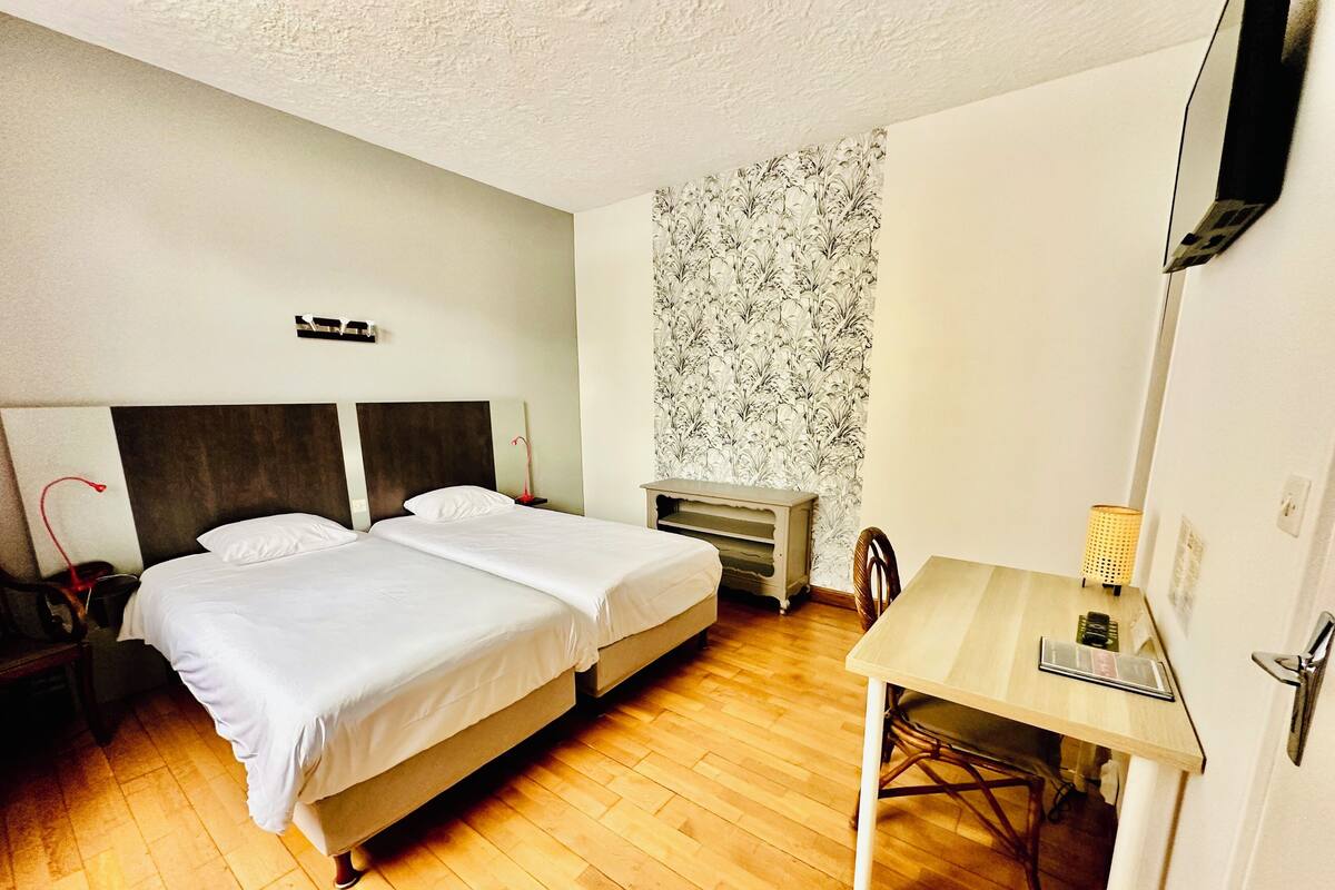 A hotel room with two beds, a wooden floor, a small table, a chair, a desk, a lamp, a TV, and a wall with a floral pattern.