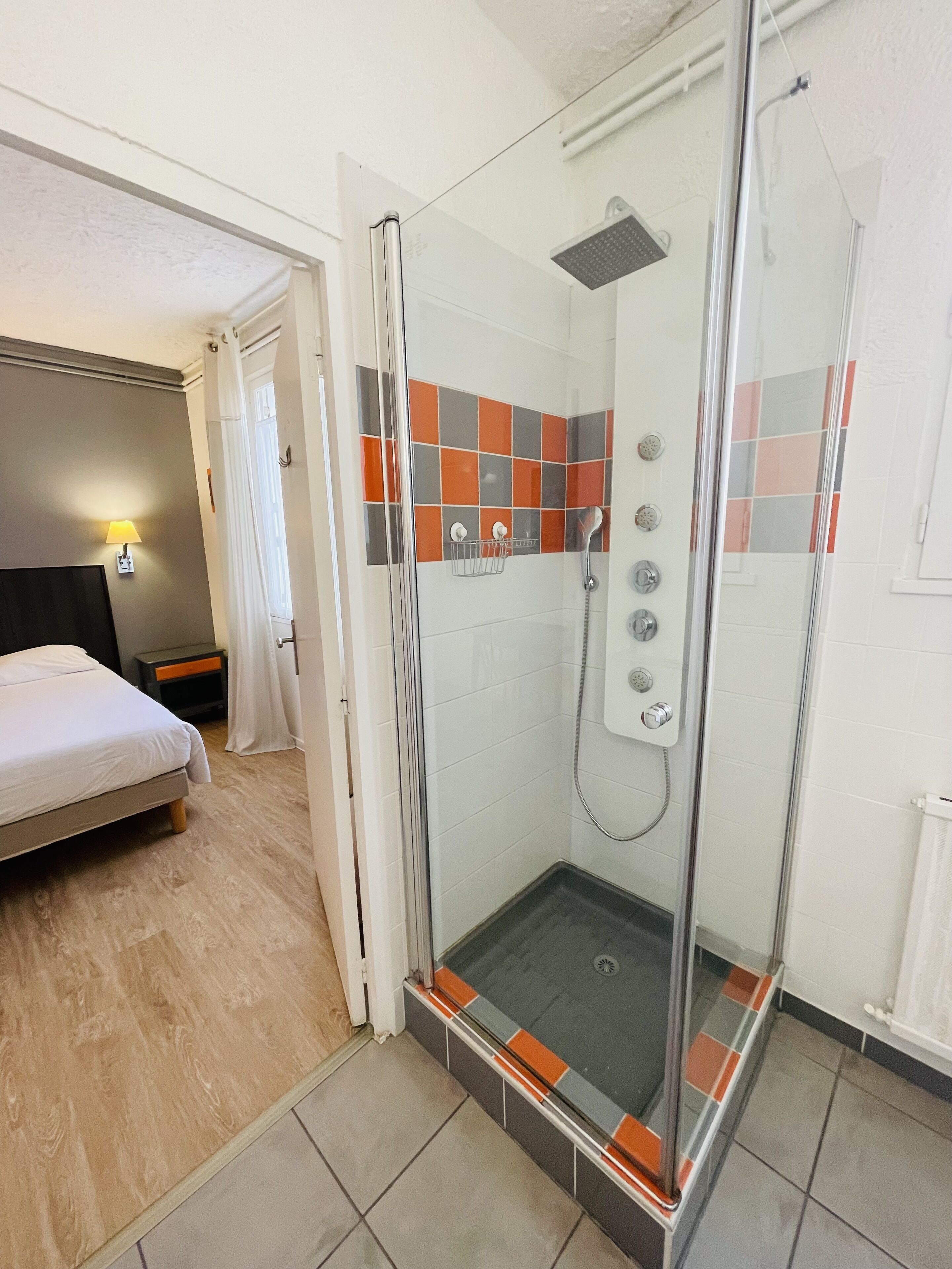 A hotel bathroom with a glass-enclosed shower, a bed, and a nightstand.