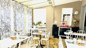 A dining area with white tables and chairs, a patterned curtain, a stone-tiled floor, and a black cabinet with kitchen items. - Hôtel Italia (Tours)