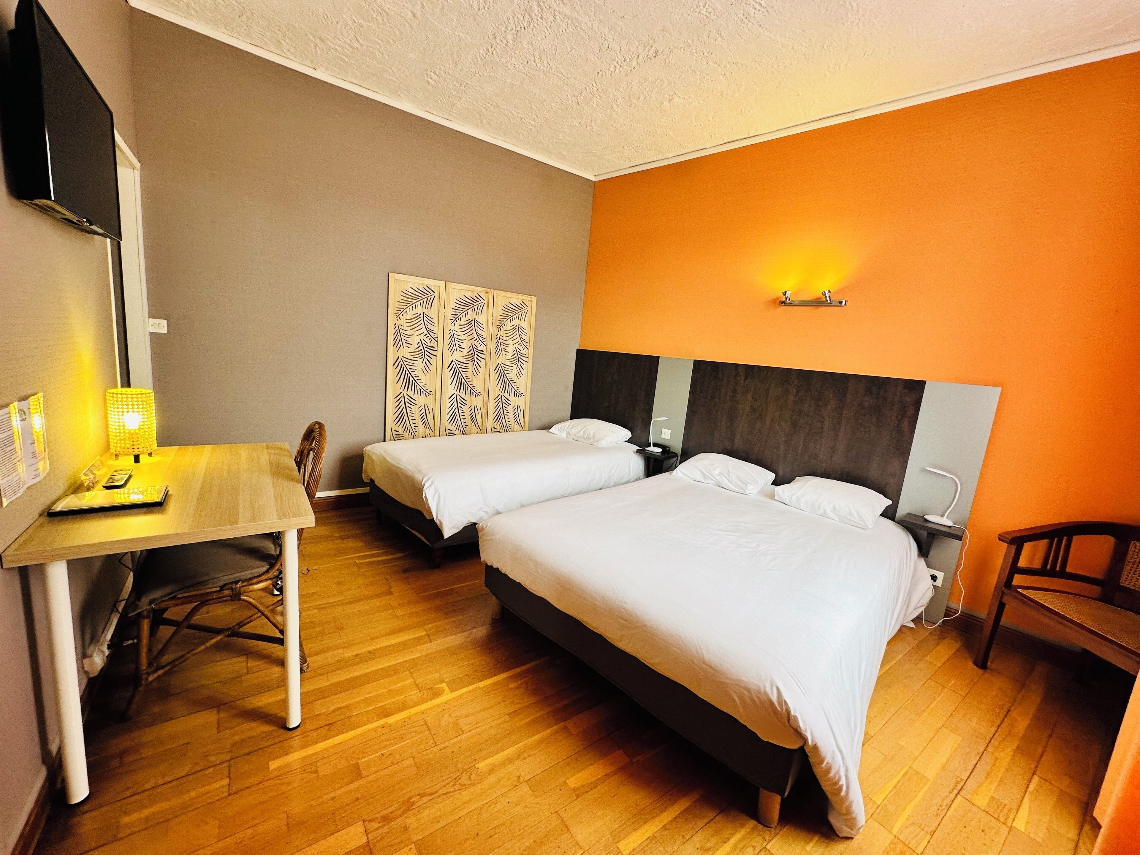 A hotel room with two beds, a desk, a chair, and a small table with a lamp.