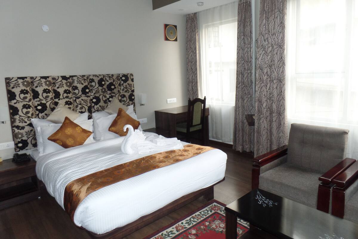 Quarto de hotel com uma cama grande, uma cadeira, uma escrivaninha e uma janela com cortinas.