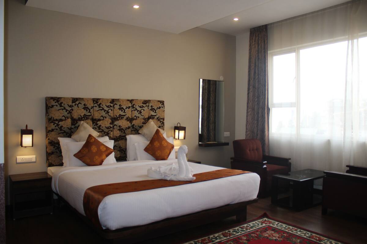 Quarto de hotel com uma cama grande, duas mesas de cabeceira, uma cadeira, uma pequena mesa e uma janela ampla com cortinas.