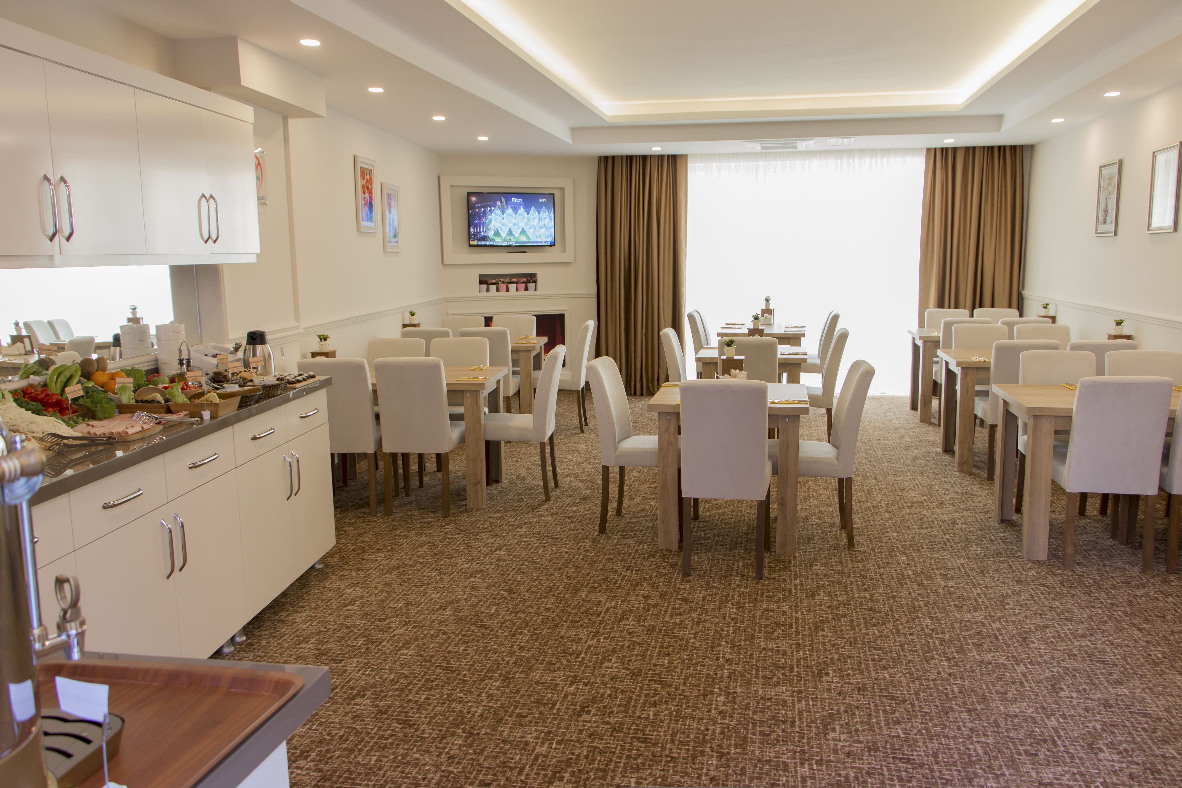 Uma área de refeições de hotel com várias mesas e cadeiras, uma televisão presa na parede e uma cozinha com armários e pia.