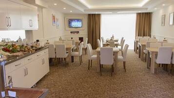 Uma área de refeições de hotel com várias mesas e cadeiras, uma televisão presa na parede e uma cozinha com armários e pia.
